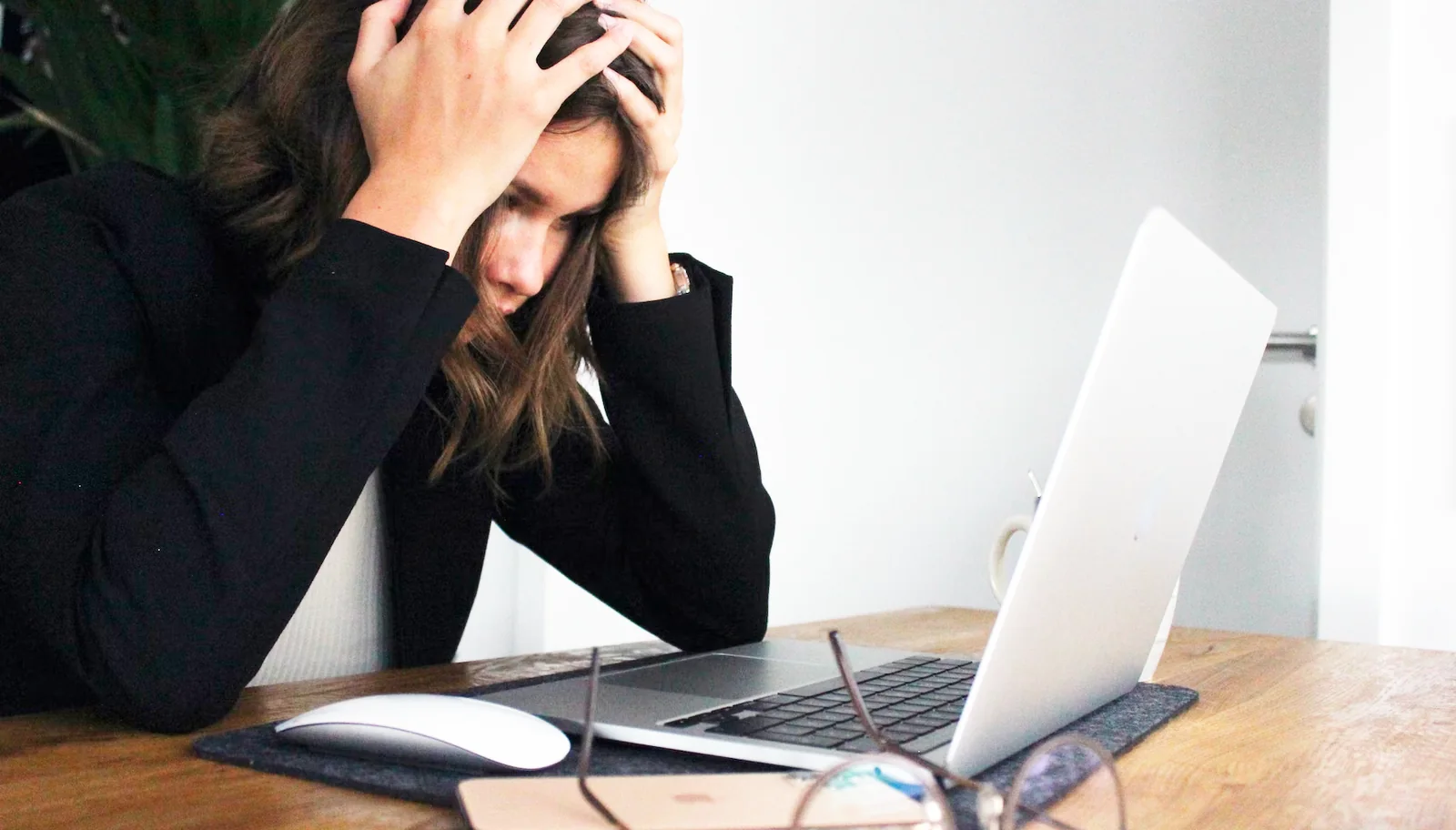 A person in a dark blazer sits at a desk with both hands on their head, looking at a laptop with glasses and a phone in front of them
