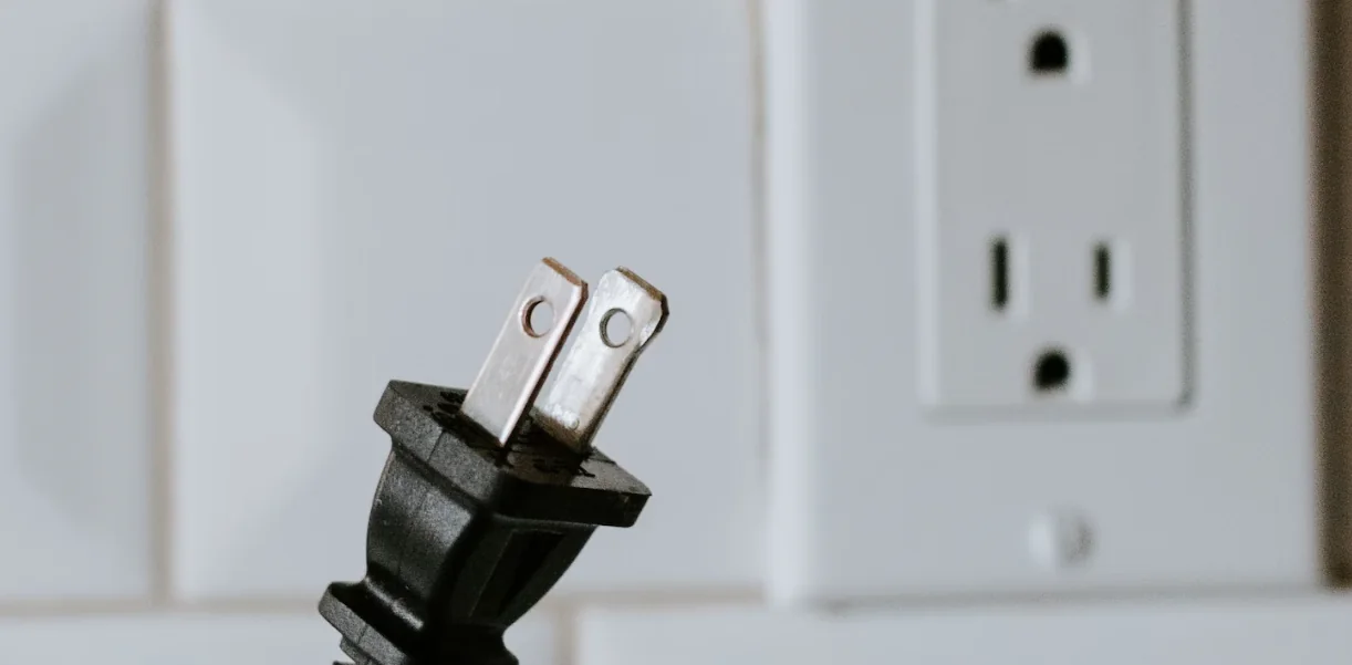 A two-prong electrical plug held in front of a wall outlet, not yet connected